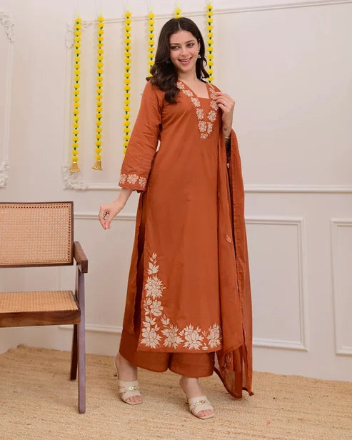 Woman wearing a rust-colored traditional outfit with white embroidery in a room with a chair and decorative wall.