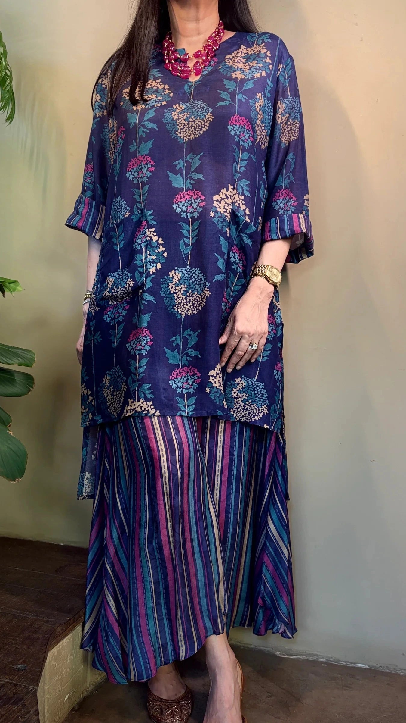 Woman wearing a blue floral dress with a striped skirt against a plain wall.