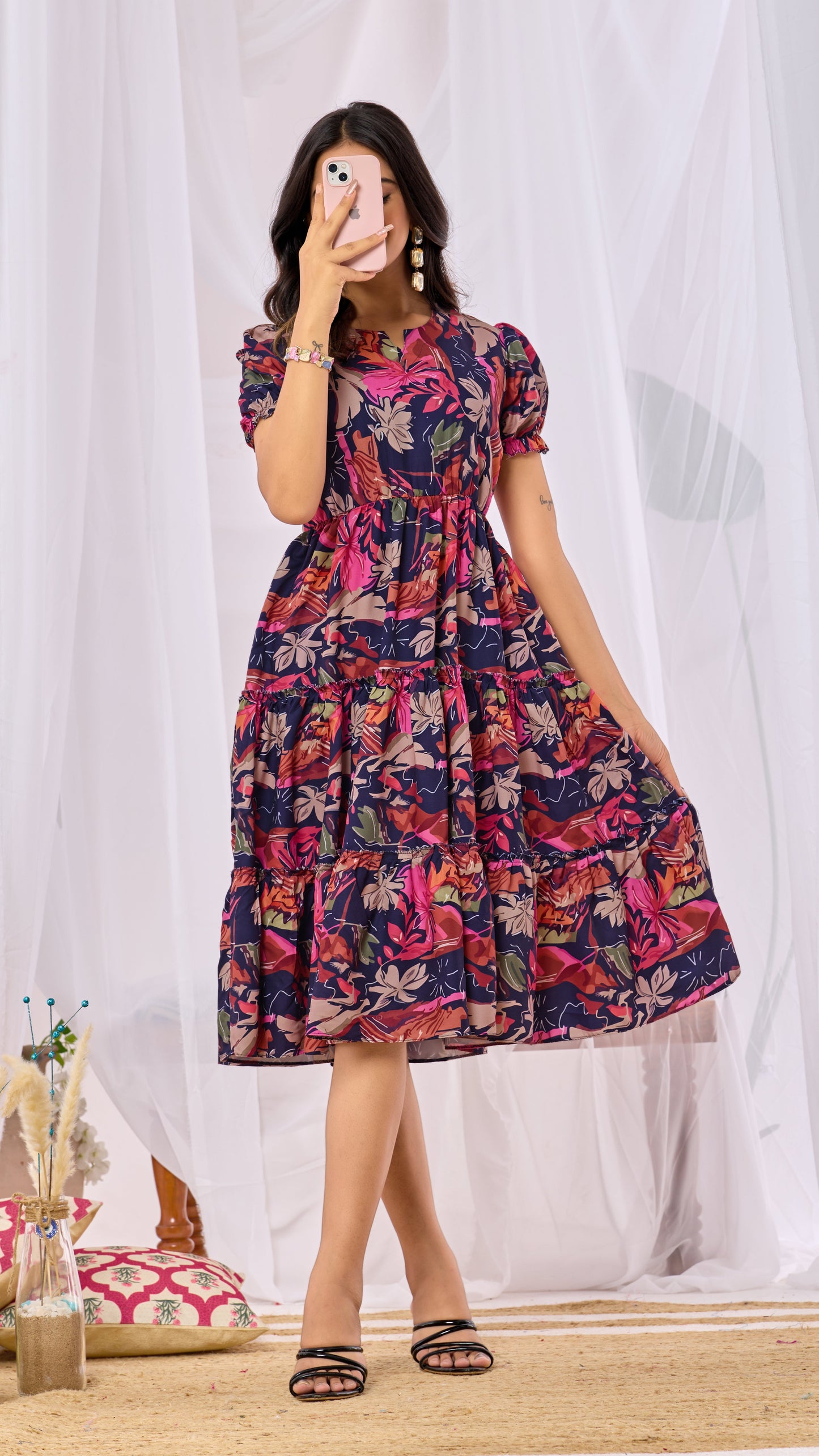 Woman wearing a colorful floral dress standing in front of white curtains.