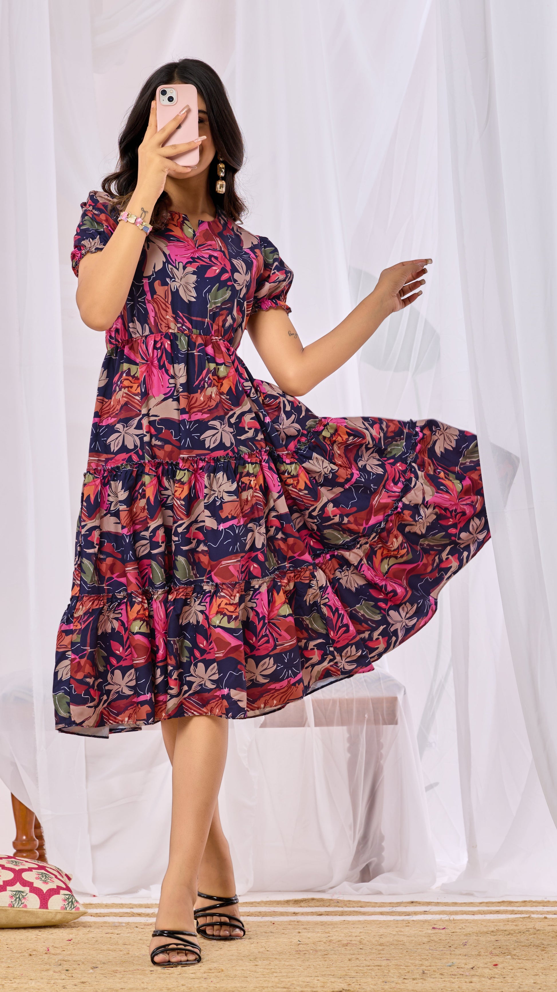 Woman wearing a colorful floral dress taking a selfie in front of white curtains.