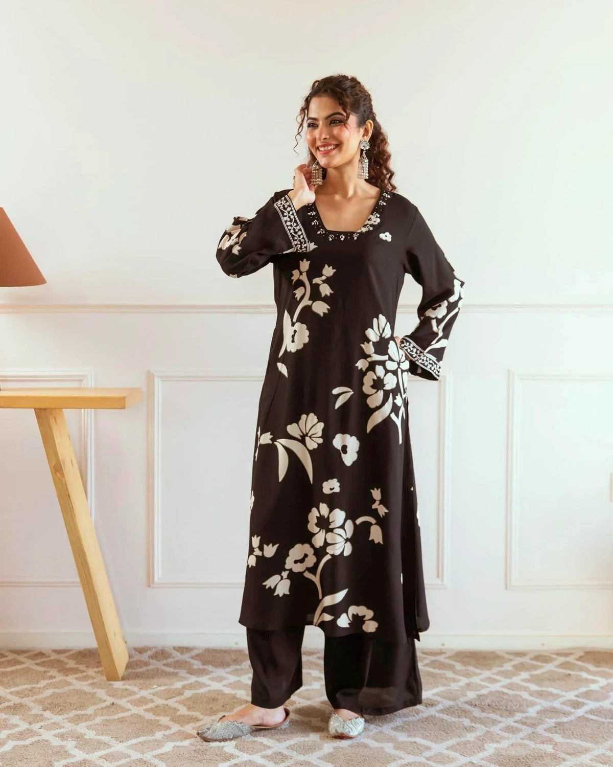 Woman wearing a black floral dress standing in a room with a white wall and wooden table.