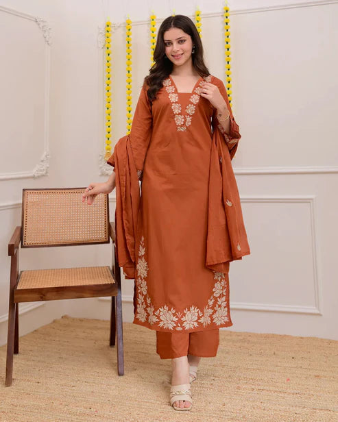 Woman wearing a rust-colored embroidered outfit standing next to a chair indoors.
