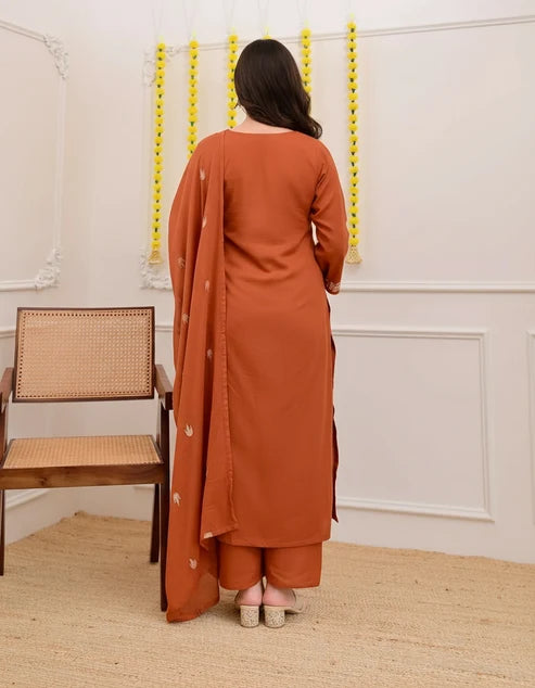 Woman in a rust-colored traditional outfit standing in a room with white walls and a wooden chair.